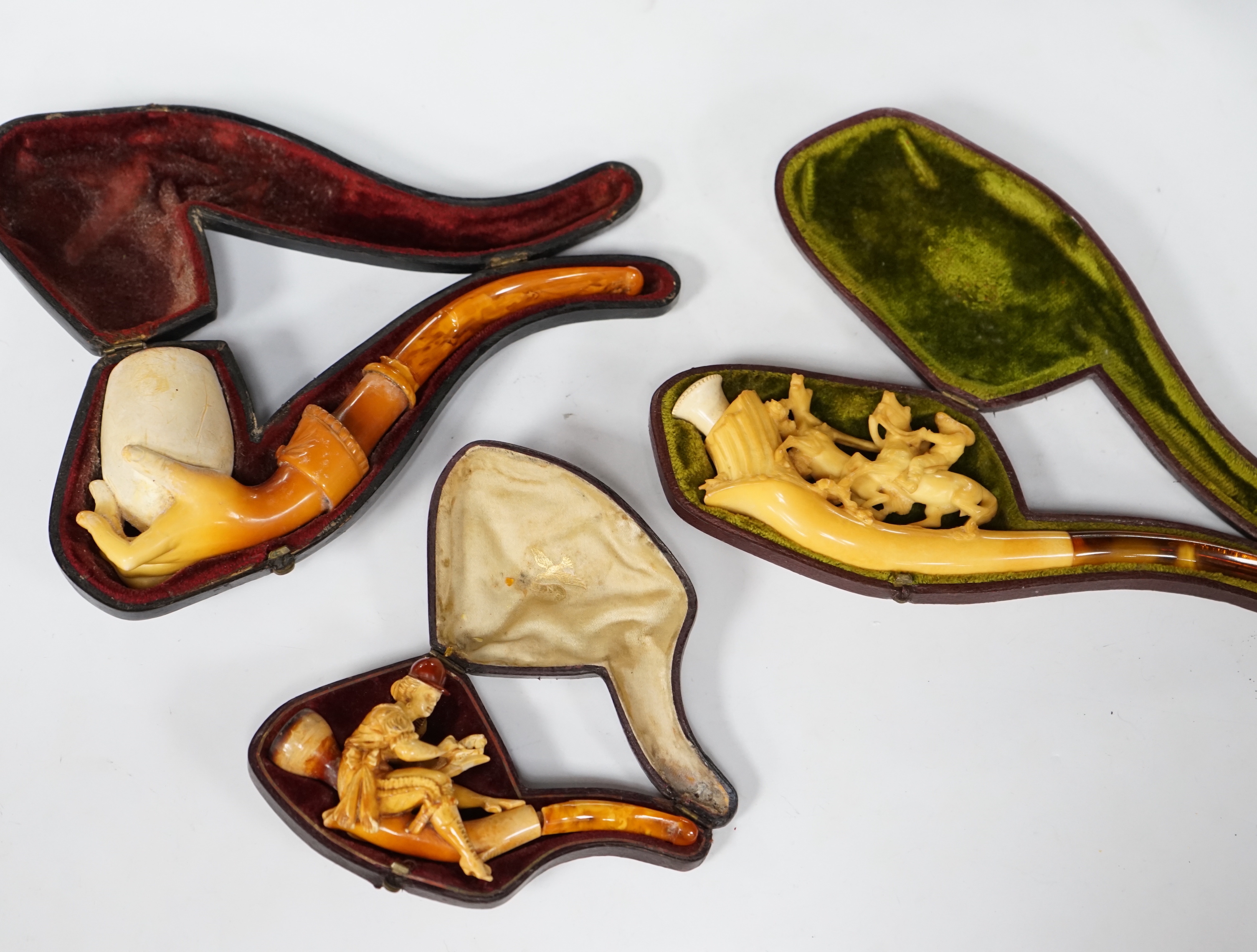 Three cased novelty Meerschaum pipes and pipe bowls, all with amber mouth pieces, one of a horses and cowboy, another a model of a hand and the other of a scantily clad lady wearing a riding cap, largest 17 cm long